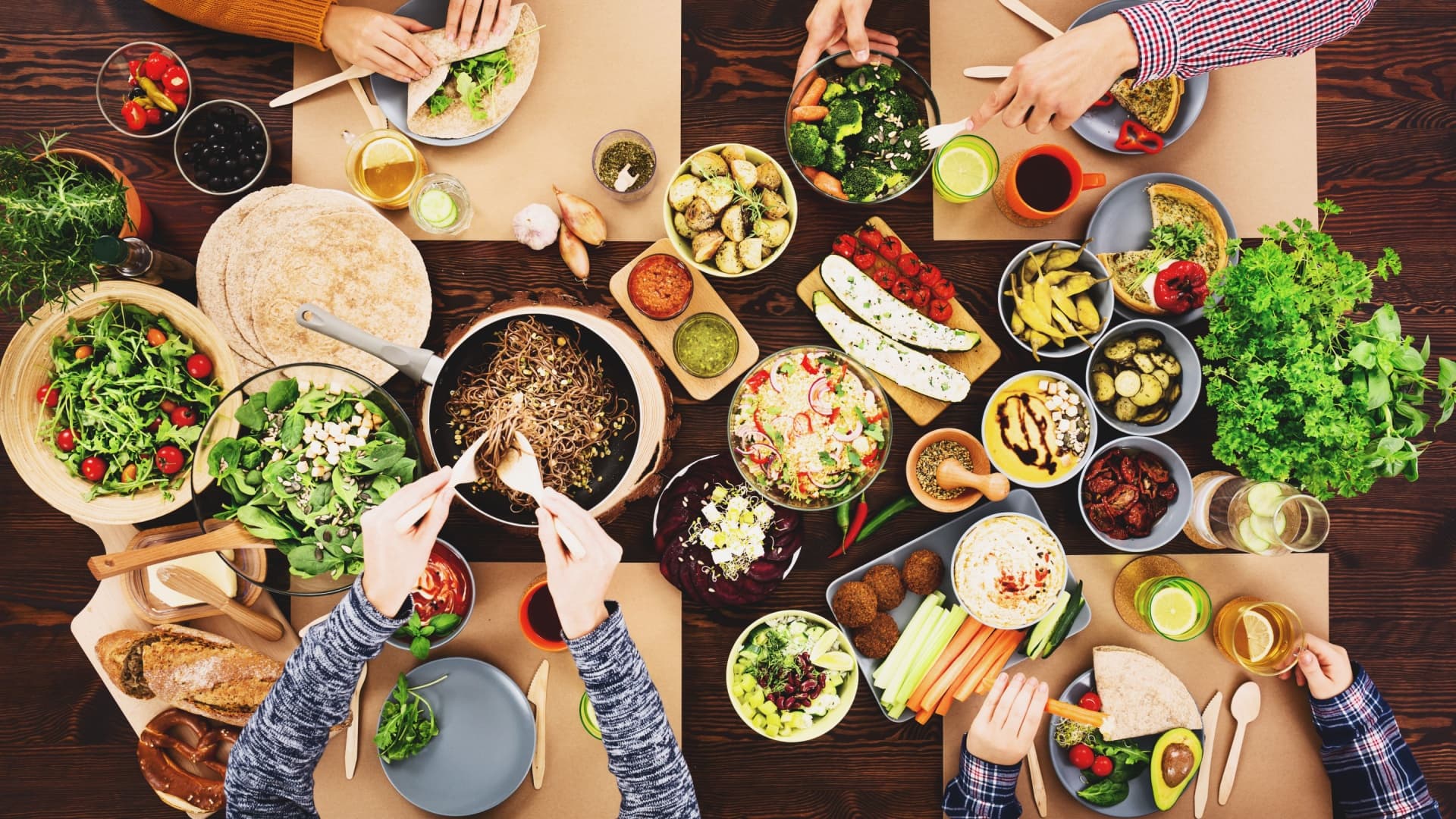 A top down view of a table laid out with many vegan dishes and salads, with 4 people taking servings to their plates using utensils