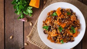 A vegan rice and beans dish with pumpkin and herbs on a white dish with garlic clove and bunch of herbs on the rustic wooden table around the dish