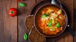 A vegan pasta dish with soup, toasted rye bread slices on a brown earthenware plate and bowl set, with whole tomatoes and pepper corn on the wooden table