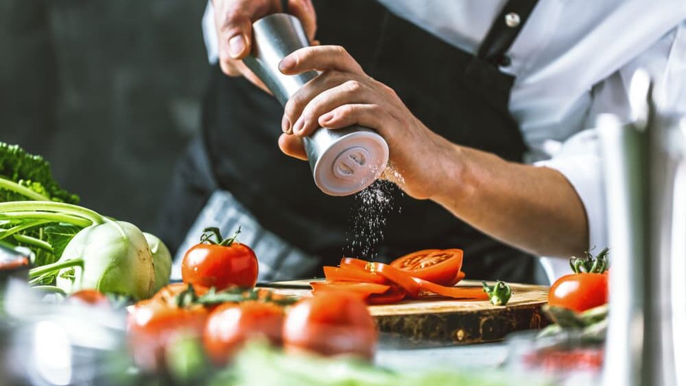 A professional chef in a commercial kitchen grinding salt onto tomato slices on a chipping board, surrounded by other fresh produce
