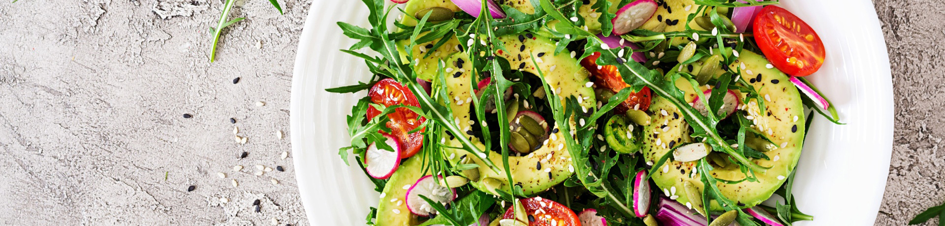 A fresh vegan salad including avocado tomato rocket online spices sesame seeds in a white dish on a stone benchtop